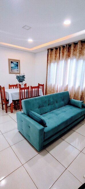 A teal sofa in a room with a dining table and chairs, white walls, and a ceiling light. - Guarujá Sol e Praia (Guarujá)