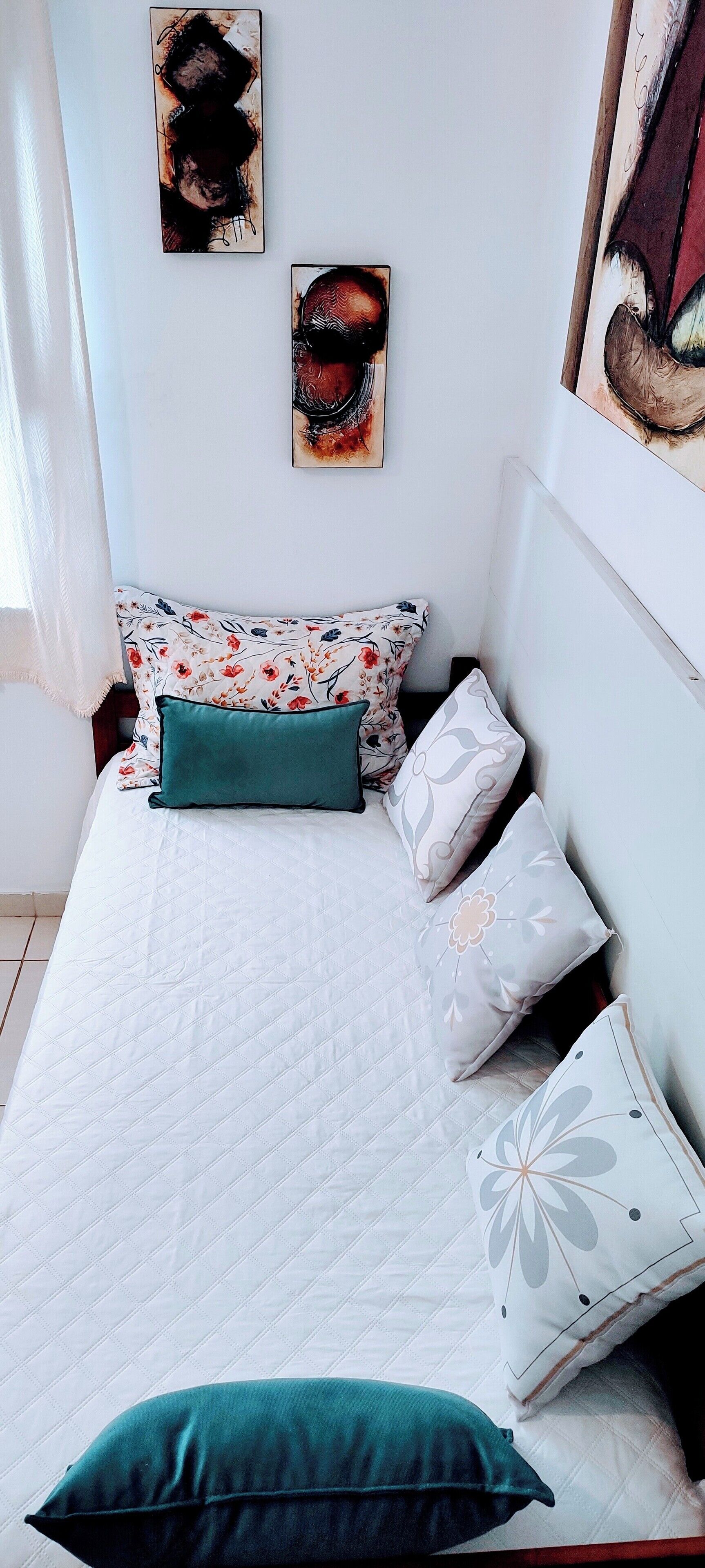 Una cama bien hecha con cubrecolcha blanco acolchado y varios cojines, incluyendo uno color turquesa. Hay dos obras de arte en la pared sobre la cama.