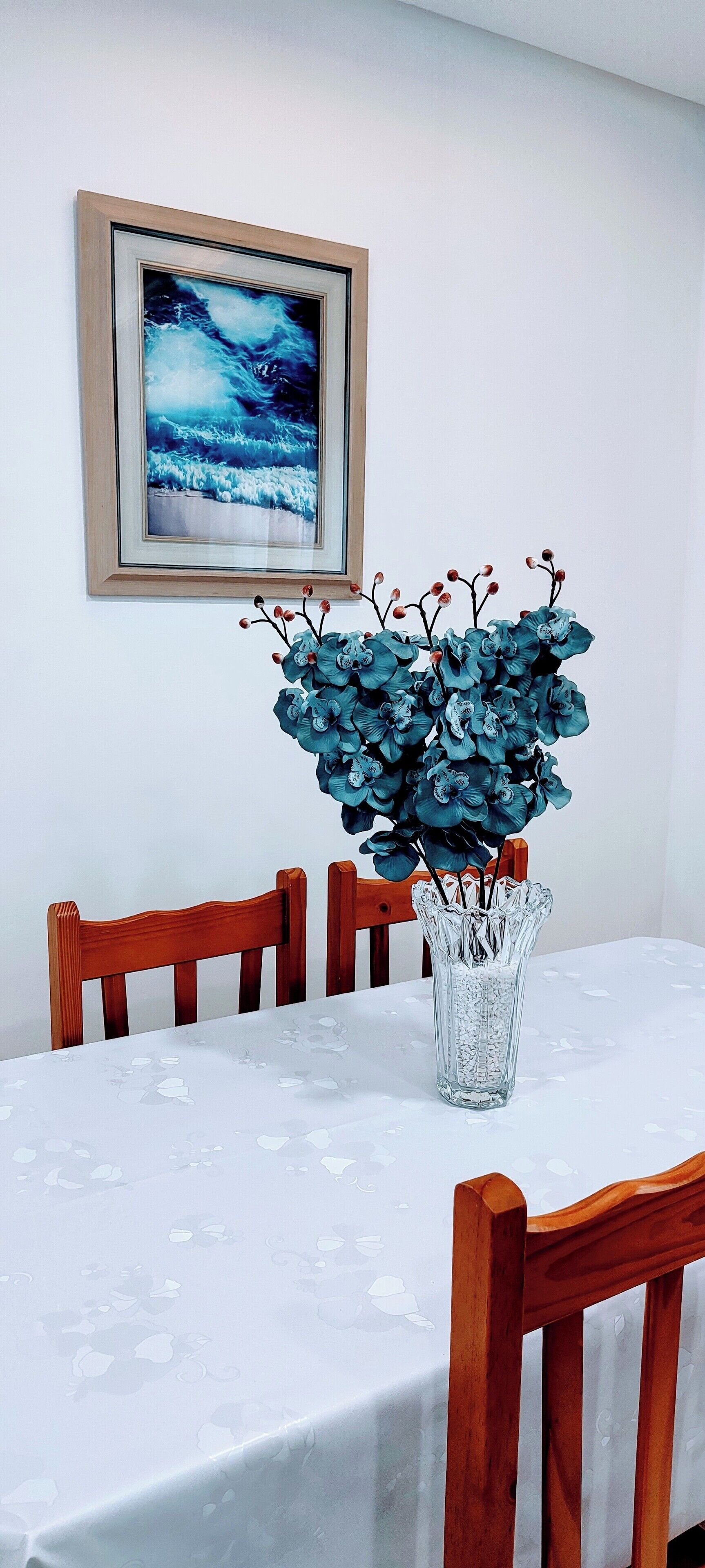 A dining table with a clear glass vase containing blue flowers, wooden chairs, and a framed artwork on the wall.
