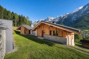 A modern wooden cabin with a stone foundation, set against a mountainous backdrop.