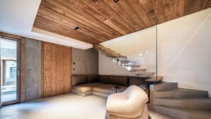 A modern interior with a wooden ceiling, a glass staircase, a sectional sofa, and a plush armchair. - Foxden - Stunning Chalet in Chamonix (Les Houches)