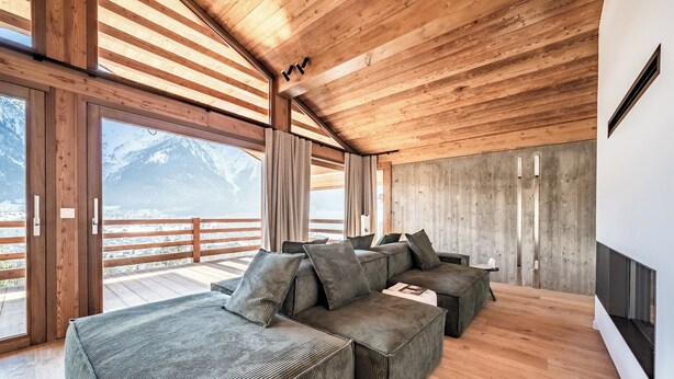 A modern living room with a large sectional sofa, wooden ceiling beams, and a view of snow-capped mountains. - Foxden - Stunning Chalet in Chamonix (Les Houches)