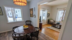 Un coin repas avec une table ronde en bois et des chaises, un saladier rempli de fruits et une grande fenêtre donnant sur une maison et un arbre.