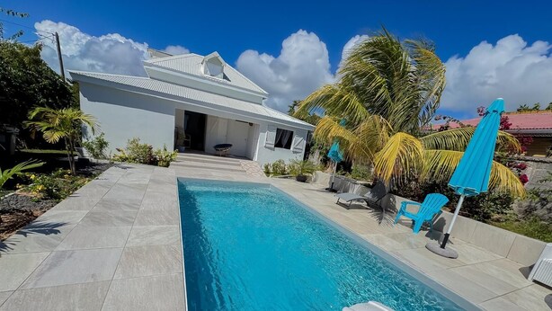 A swimming pool with a slide, surrounded by a patio with chairs and umbrellas, and a white building in the background. - Villa Fleur de Sel with swimming pool 5 minutes walk from the beach (le gosier)
