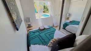 A compact bedroom with a bed, a bedside table, a lamp, and a window with a view of a pool. - Villa Fleur de Sel with swimming pool 5 minutes walk from the beach (le gosier)