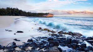 Ein Strand mit felsigem Vordergrund, einem Sandstrand und einer großen Felsformation im Hintergrund.
