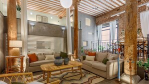 A spacious living area with a high ceiling, exposed wooden beams, and a large window with blinds. There are two sofas, a coffee table, and wicker chairs. - Historic Loft for 7 in Heart of Madrid (Madrid)