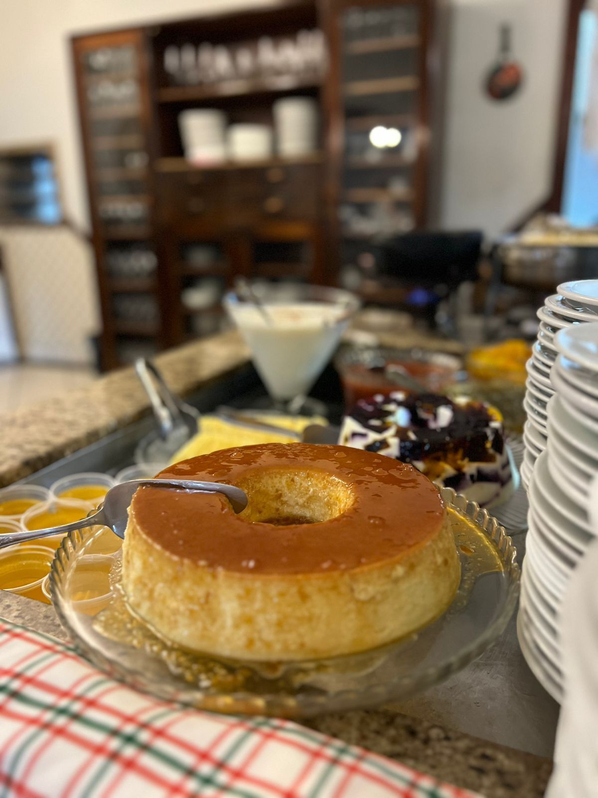 Una presentación de postres con un flan de caramelo sobre un plato de vidrio, rodeada de diversos dulces y una pila de platos blancos.