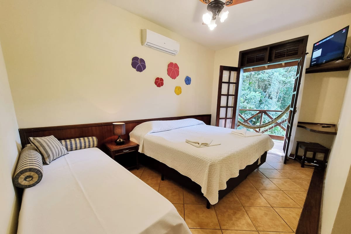 Quarto de hotel com duas camas, mesa de cabeceira com abajur, ar condicionado, televisão e varanda com vista para o verde.