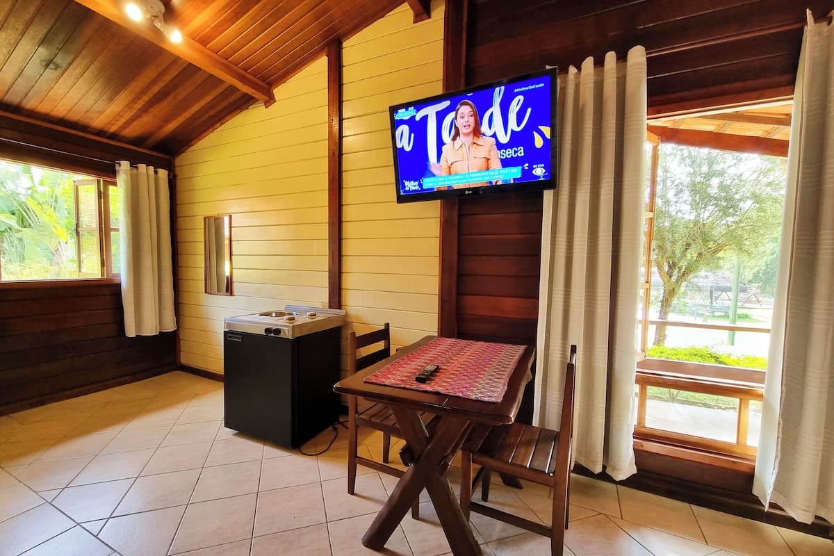 Um quarto aconchegante com teto de madeira, televisão fixada na parede, mesa pequena com toalha de mesa estampada e janela com cortinas.