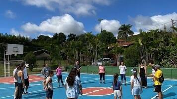 A group of people on a blue outdoor basketball court with a basketball hoop, surrounded by greenery and a partly cloudy sky.