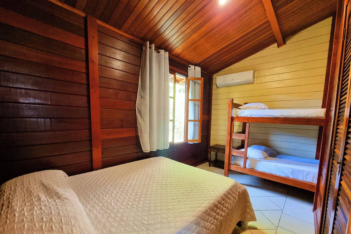 Quarto de cabana de madeira com beliche, uma cama de solteiro, paredes de madeira, janela com cortinas e um aparelho de ar condicionado.