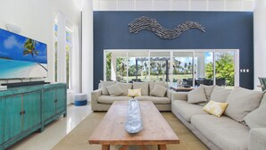 A living room with a large TV, a wooden coffee table, a teal cabinet, a beige sofa, and a view of the outdoors through large windows. - Golf Villa in Bavaro - Pool, Jacuzzi, Staff (Punta Cana)