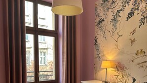 Een kamer met een houten vloer, een witte tafel, een groene bank, een witte dressoir, een hanglamp, een raam met gordijnen en bloemenbehang.