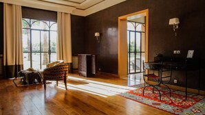 A spacious room with wooden flooring, a large window with curtains, a patterned armchair, a desk with a chair, and a decorative rug.