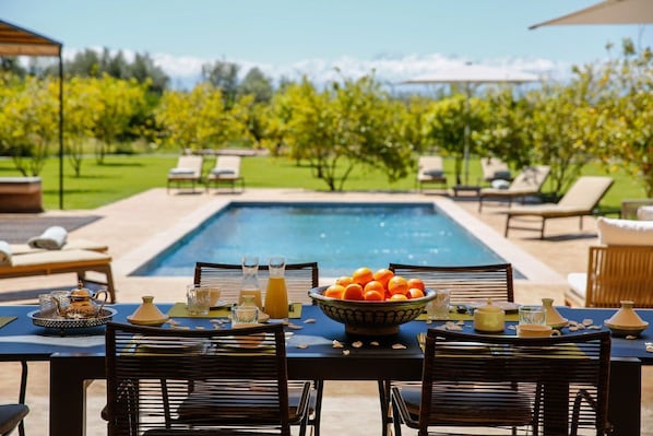 An outdoor dining area with a pool, lounge chairs, and a table set for breakfast with a bowl of oranges.
