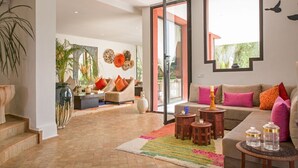 Uma sala de estar ampla com um sofá, mesa de centro e almofadas decorativas. Há uma janela grande que proporciona luz natural e vista para o exterior.