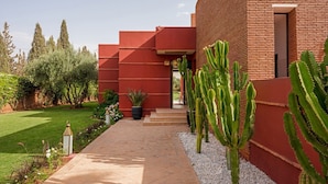 Um edifício moderno com fachada vermelha, um caminho pavimentado e cactos no jardim.