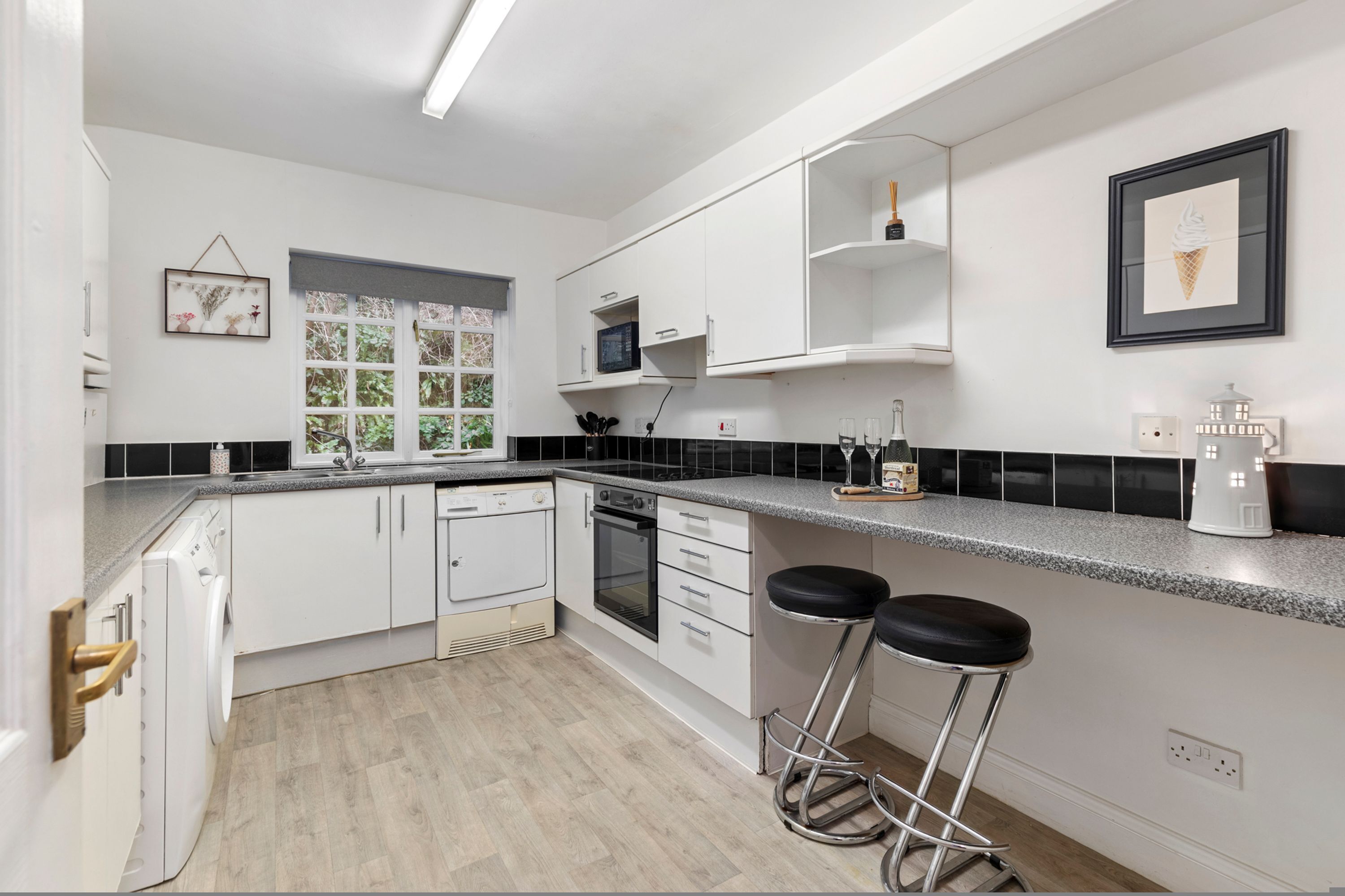 Une cuisine moderne avec des meubles blancs, un comptoir noir et une fenêtre donnant sur un espace verdoyant. On y trouve deux tabourets de bar et une petite étagère ornée d’objets décoratifs.
