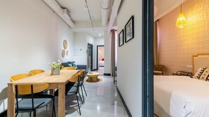A modern hotel room with a wooden dining table, chairs, a bed, and a hallway leading to another room.