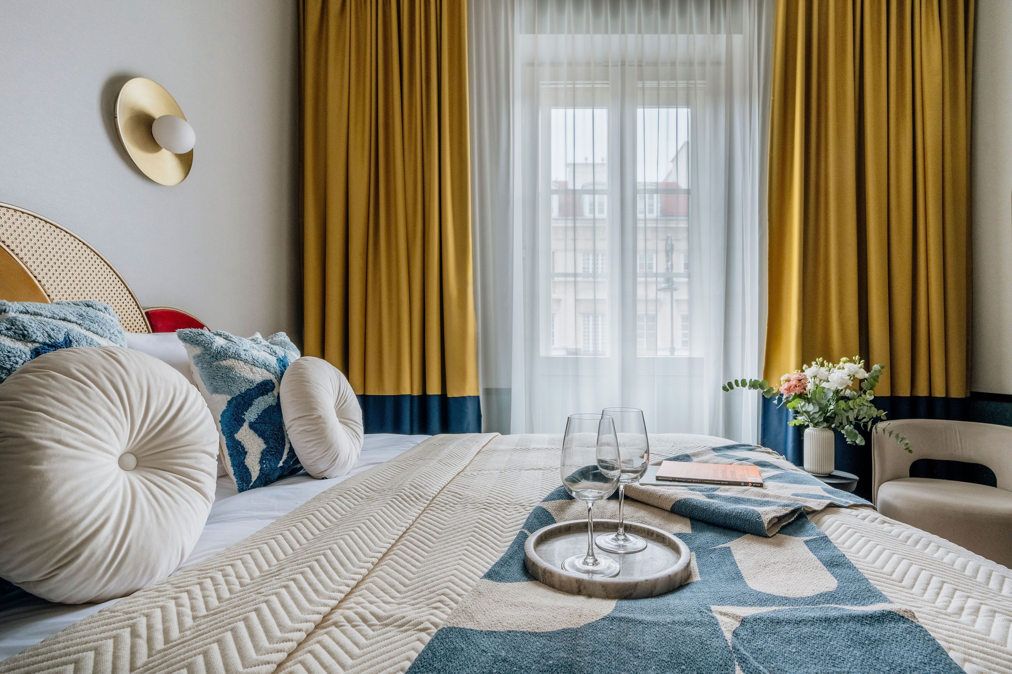 A hotel room with a bed, a bedside table with two wine glasses, a vase with flowers, and a view of buildings through sheer curtains.