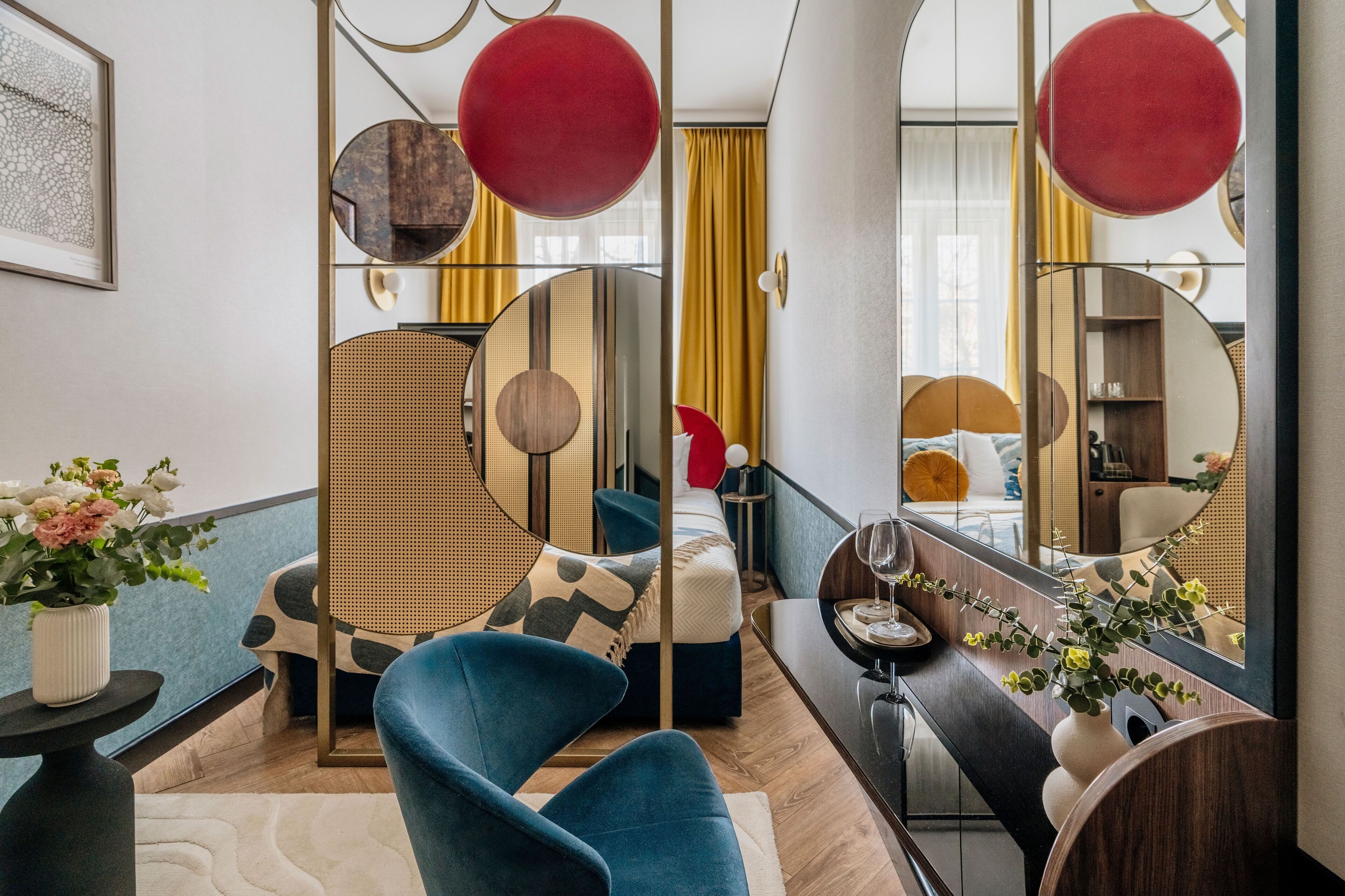 A modern hotel room with a bed, a blue armchair, a round mirror, and a small table with a vase of flowers.
