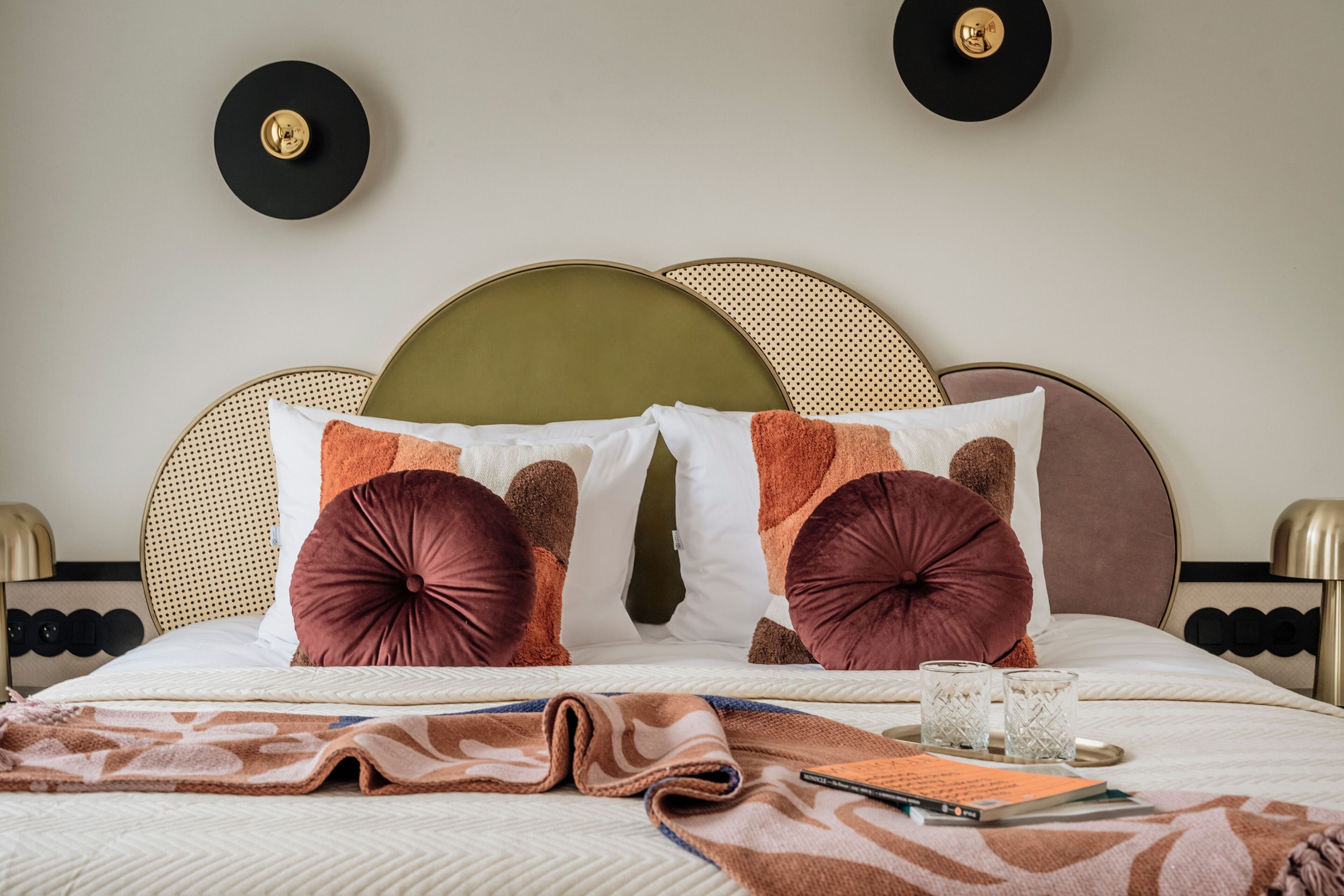 Cama bem arrumada com roupa de cama estampada, duas relógios fixados na parede e uma mesa de cabeceira com abajur.