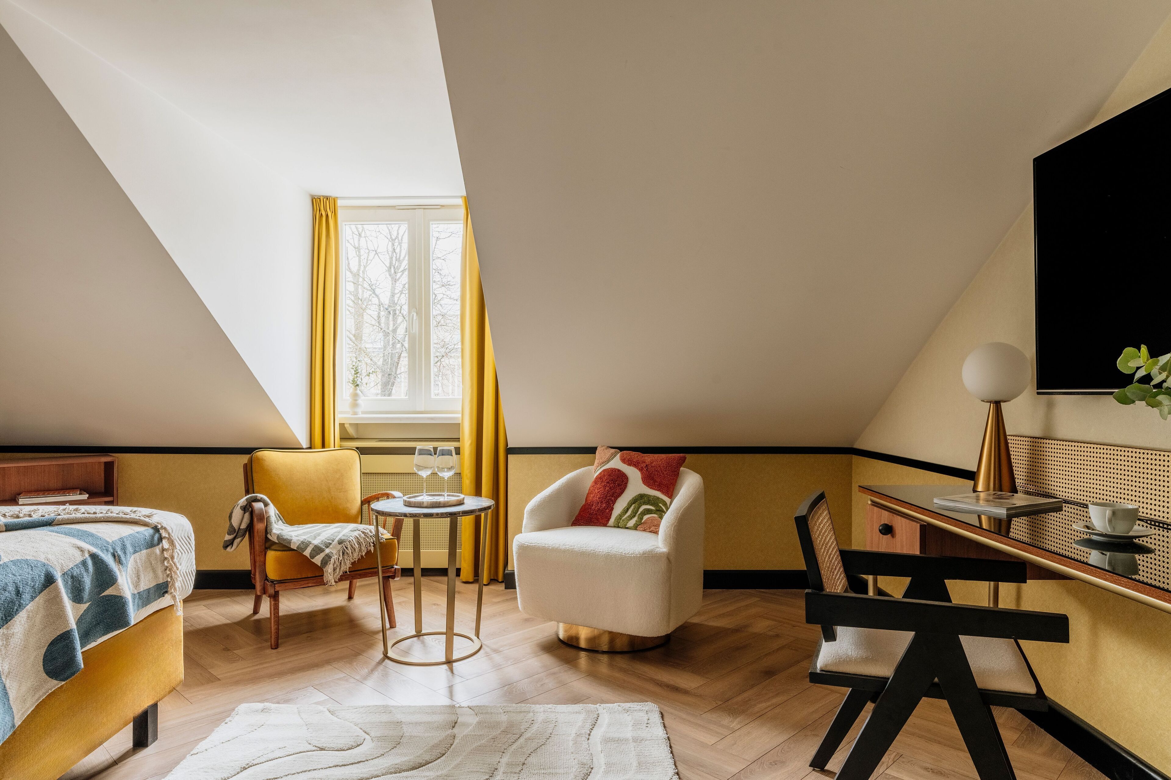 A modern hotel room with a bed, a yellow armchair, a white armchair, a small table with a lamp, and a TV mounted on the wall.