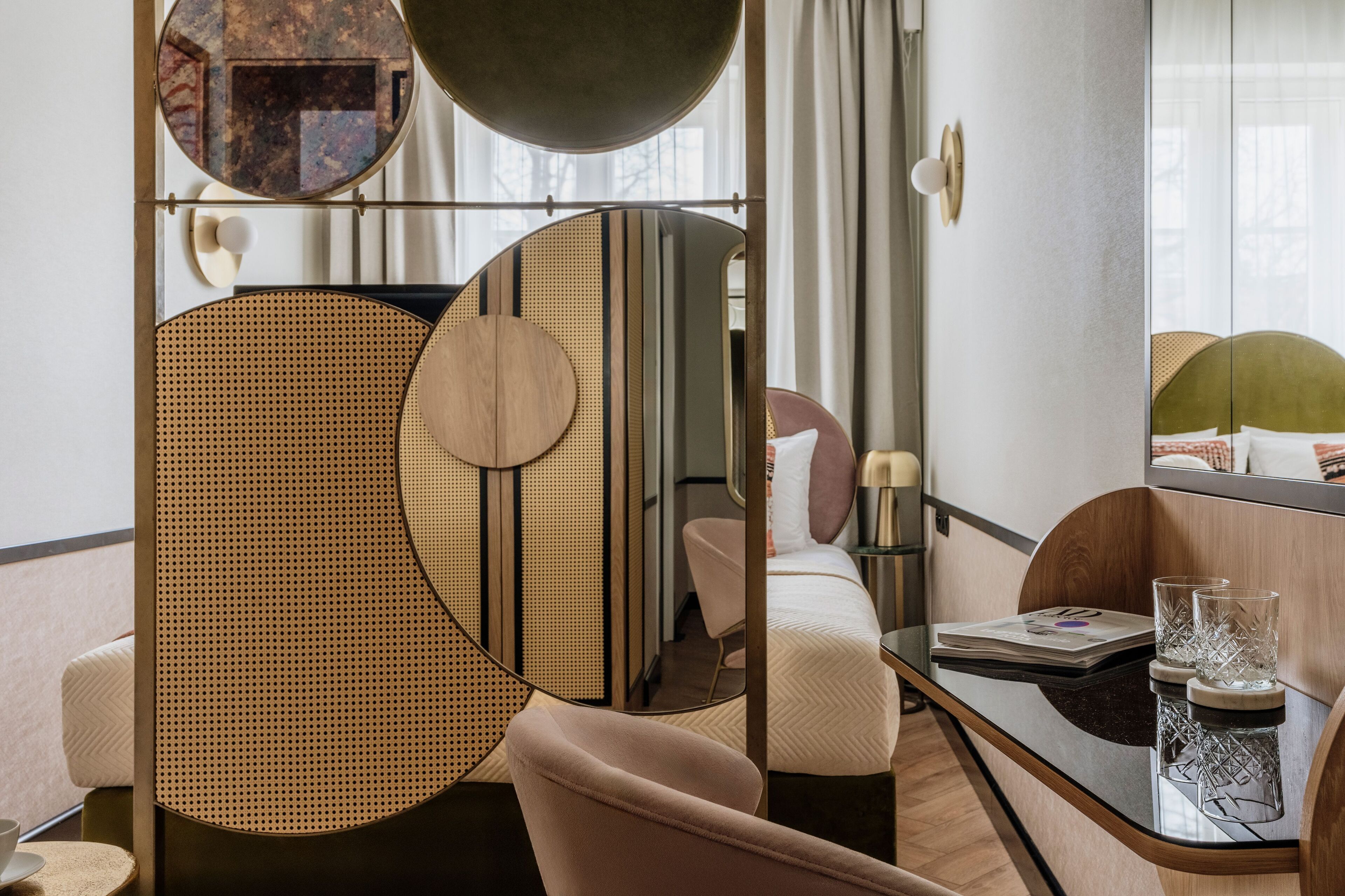 Una habitación de hotel moderna con una cama, un espejo redondo, una mesa redonda con superficie de vidrio y una mesita de noche con lámpara.