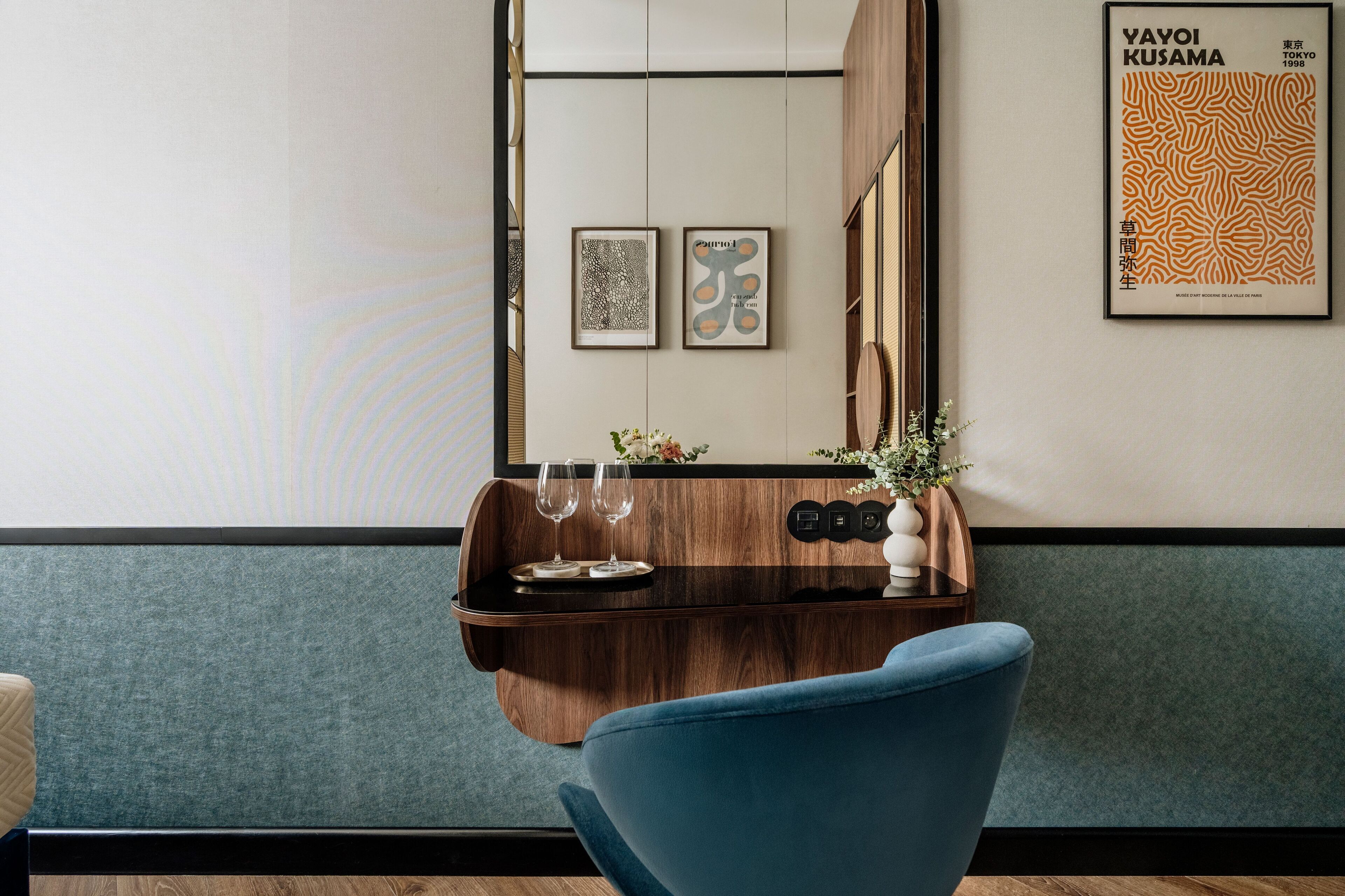 A modern hotel room with a wooden console table, two framed artworks, a mirror, and a blue chair.