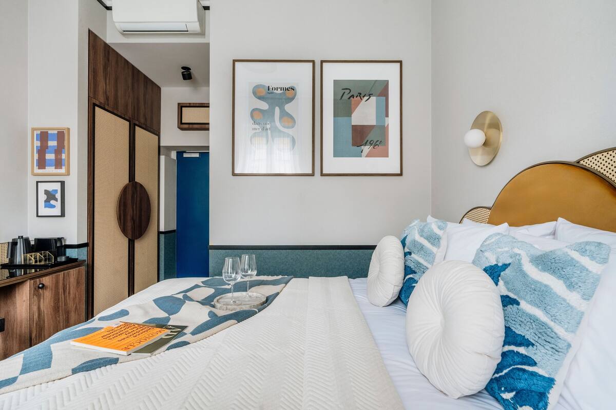 Une chambre d’hôtel bien rangée, avec un lit, des oreillers, une table de chevet sur laquelle se trouvent un livre et des lunettes, et deux œuvres d’art encadrées accrochées au mur.
