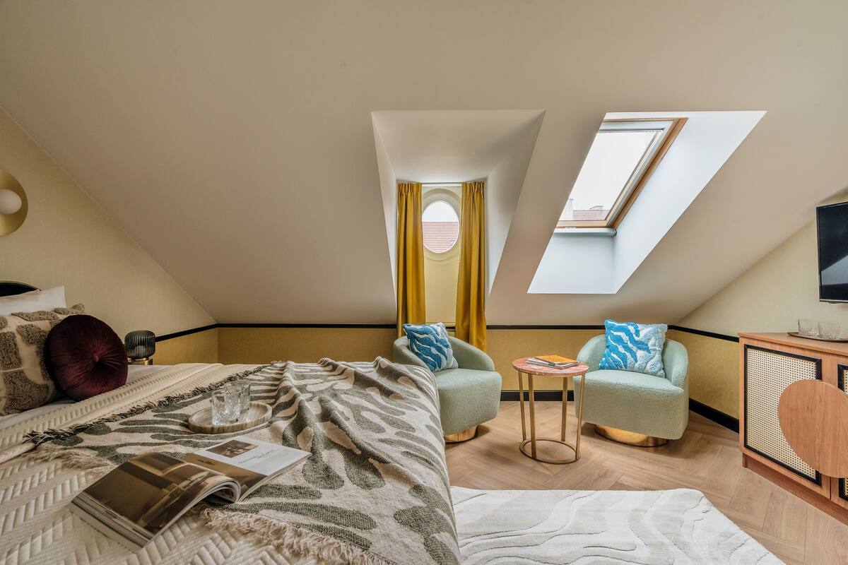 Une chambre à coucher avec un lit, une petite table, deux fauteuils, un téléviseur et une lucarne.