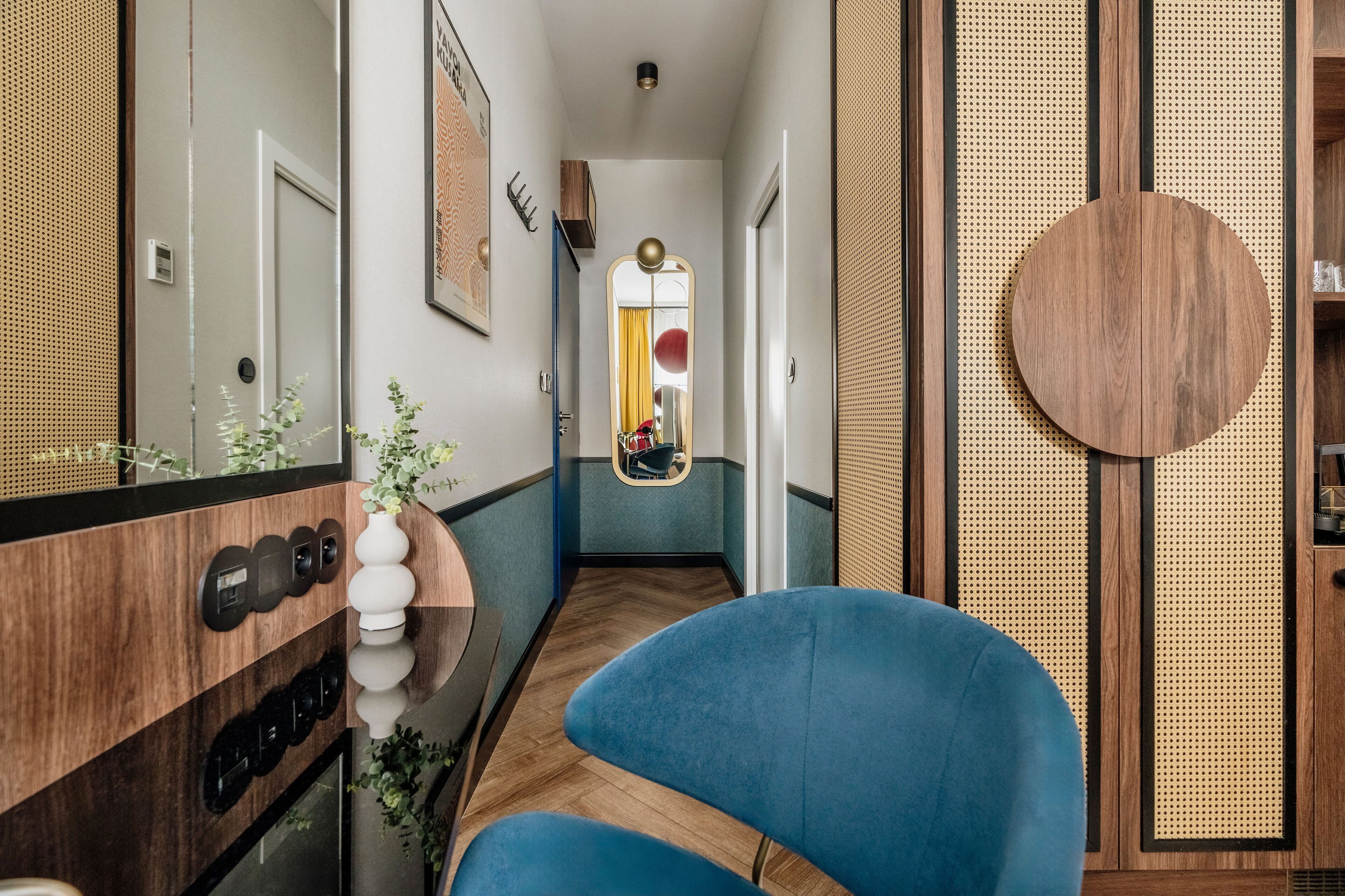 A narrow hallway with a blue chair, a mirror, and a round wooden panel on the wall.