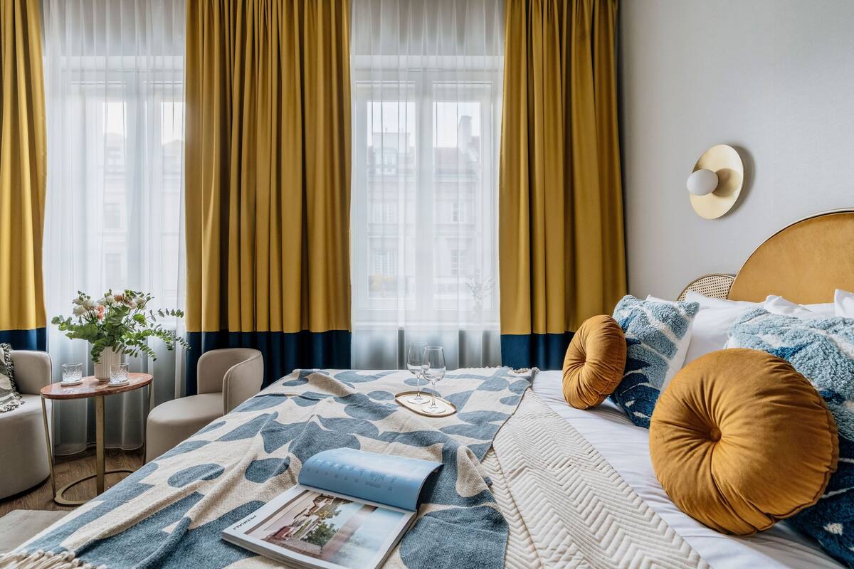 Une chambre d’hôtel avec un lit, une petite table sur laquelle est posé un vase de fleurs, et une fenêtre avec des rideaux.