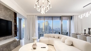 A modern living room with a large sectional sofa, a flat-screen TV, and a city view through floor-to-ceiling windows. - Elegant 2B at Boulevard Pointburj & Fountain View (Dubai)