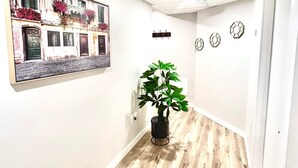 A hallway with a wooden floor, a potted plant, a wall-mounted clock, and a framed picture of a building.