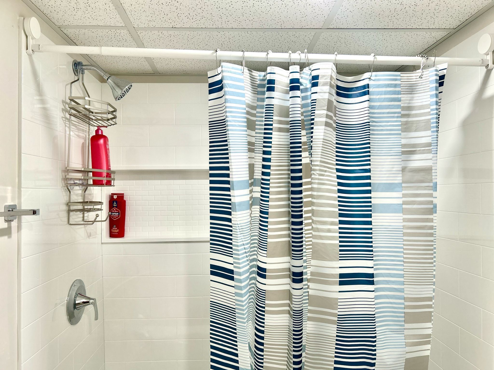 Une salle de bain avec des murs recouverts de carreaux blancs, une douche avec un pommeau de douche amovible, un flacon de shampoing rouge et un rideau de douche à rayures.