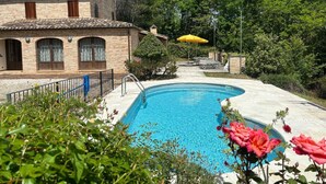 Una piscina con borde de baldosas, rodeada de abundante vegetación y un edificio de piedra con ventanas arqueadas.