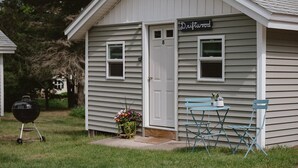 A small, single-story building with a 'Driftwood' sign, a door, two windows, a small porch with blue chairs, and a barbecue grill in the yard. - Brookside Cottages and Resort Driftwood Cottage (Grawn)