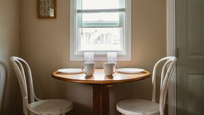 A dining area with a round wooden table, two white chairs, and a window with blinds. - Brookside Cottages and Resort Driftwood Cottage (Grawn)