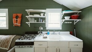 A compact kitchen with a gas stove, white cabinets, and a sink. There are shelves above the stove holding dishes and glasses. - Brookside Cottages and Resort Willow Cottage (Grawn)