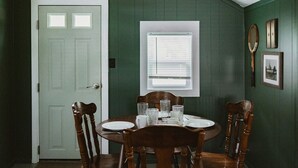 A dining area with a round table, four wooden chairs, a white door, and a window with blinds. - Brookside Cottages and Resort Willow Cottage (Grawn)