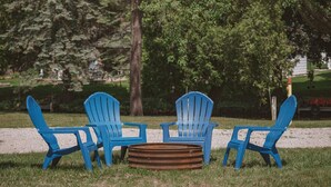 A group of four blue Adirondack chairs arranged around a circular fire pit in a grassy park area with trees in the background. - Brookside Cottages and Resort Willow Cottage (Grawn)
