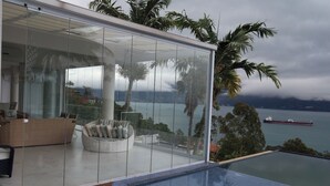 Una piscina infinita moderna con vistas al mar y un barco grande. En el interior, hay una zona de estar con paredes de cristal, un banco acolchado y suelo de madera.