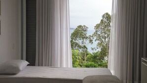 Un dormitorio con cama, cortinas blancas y vistas a los árboles y al mar.