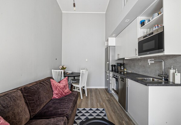 A compact kitchen with a dining area, a brown sofa with a pink pillow, and a small table with chairs. - Gesta Attefall in Gesta (Farsta)