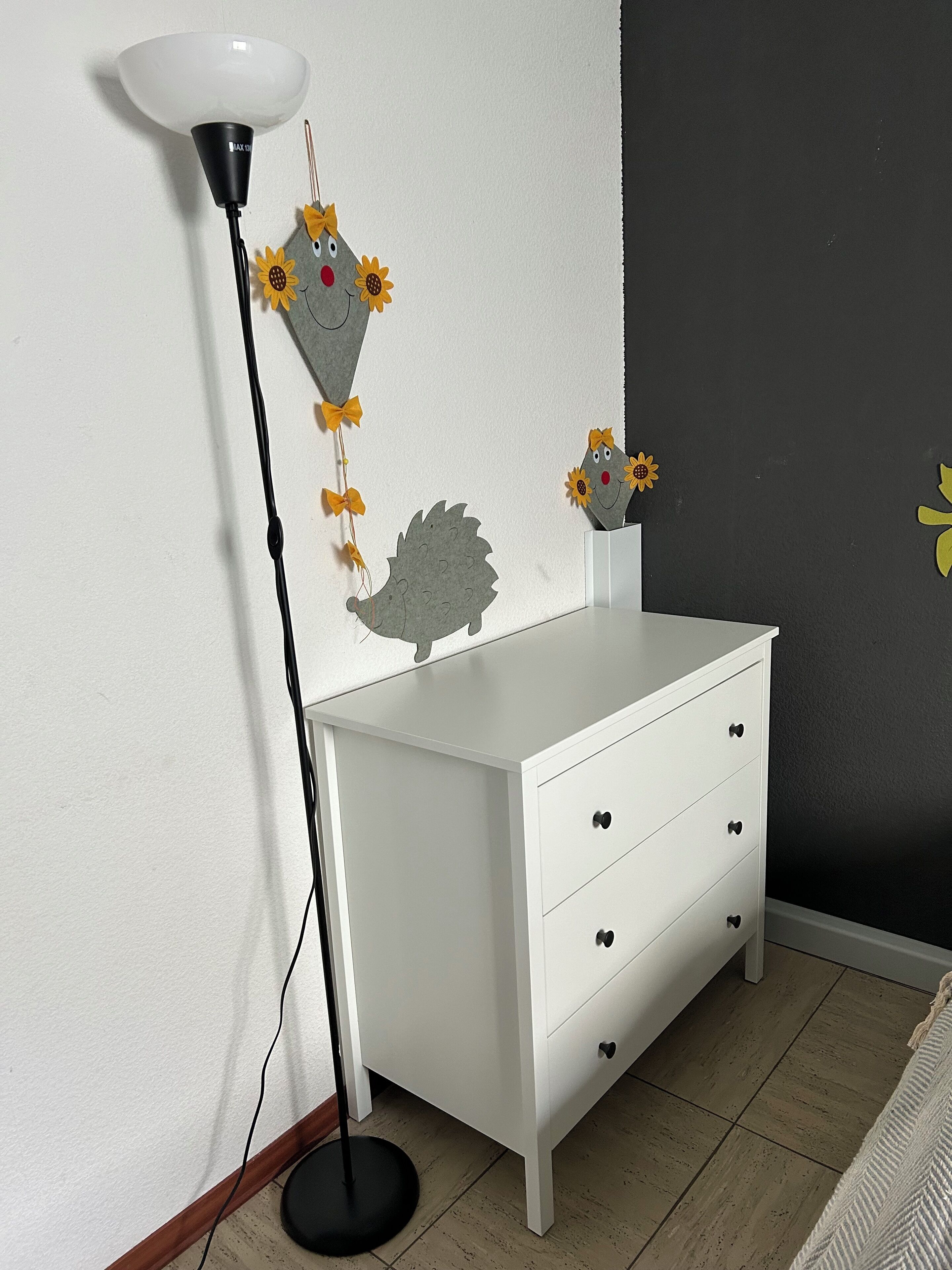 Ein weißer Kommode mit drei Schubladen, eine schwarze Stehlampe und Wandkunst mit einem Igel und Sonnenblumen.