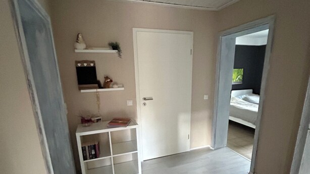 A hallway with a white door, a small shelf with decorative items, and a glimpse of a bed in the adjacent room. - Cozy vacation apartment directly at the highway entrance (Freudenberg)