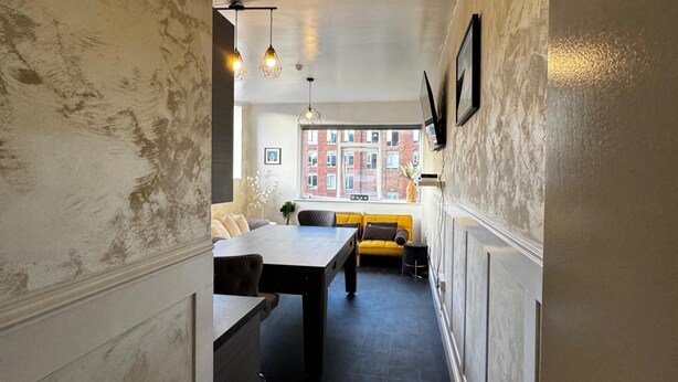 Un couloir étroit avec une table et des chaises de salle à manger, un canapé jaune et une grande fenêtre donnant sur des immeubles.