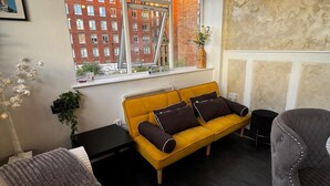 Une pièce avec un canapé jaune, un fauteuil gris, un radiateur, une fenêtre donnant sur des immeubles en briques rouges et un tableau encadré accroché au mur.